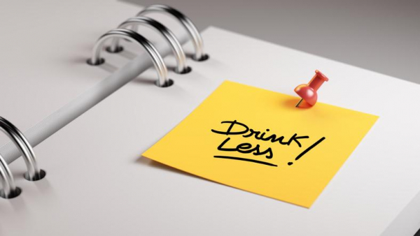 White notebook with a yellow sticky note that says Drink Less! pinned to the page by a red thumbtack.